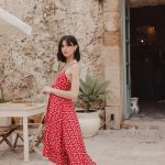 "Woman wearing a sleeveless red maxi dress with a white diamond pattern, walking outdoors in a rustic courtyard."