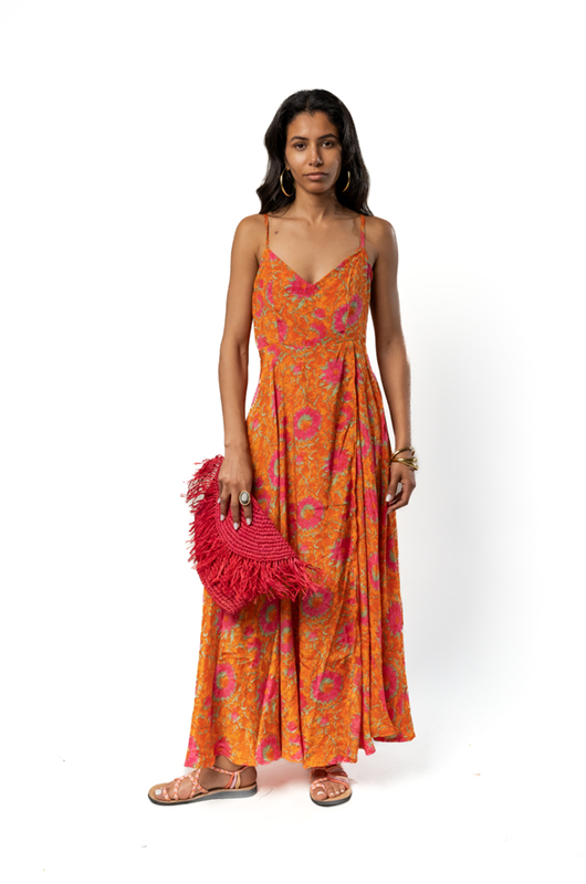 Woman in a flowing orange maxi dress with pink floral patterns, holding a fringed red clutch, standing against a white background."