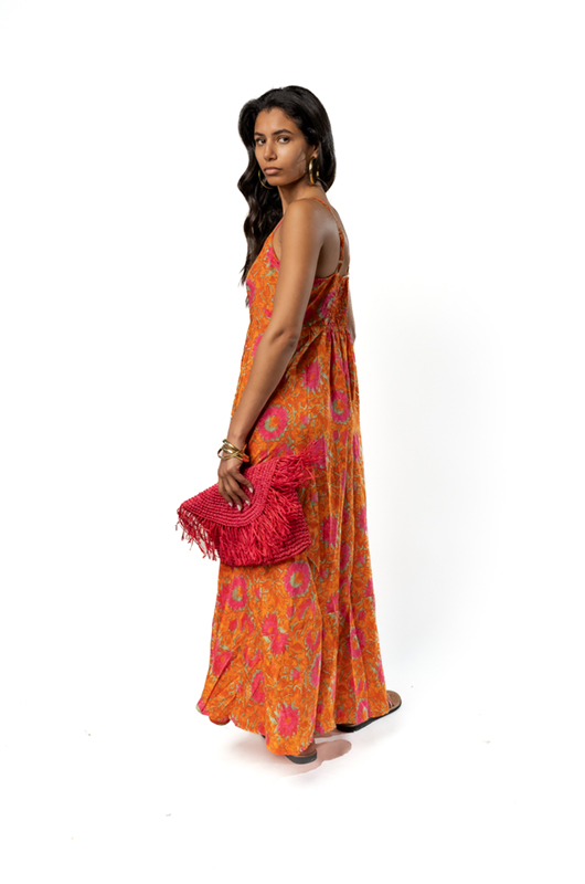 "Back angled view of a woman in a sleeveless orange maxi dress with pink floral print, holding a red fringed clutch."
