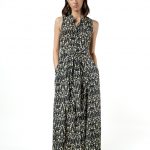 A woman wearing a sleeveless, full-length black and beige ikat print dress with a button-down front and waist tie, standing with her hands in the pockets, front view.