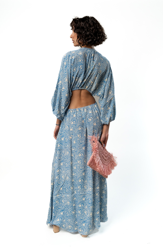A model shows the back of a blue patterned maxi dress with open back and cut-out detailing, tied at the waist with a ring in the front, while holding a pink fringed clutch.