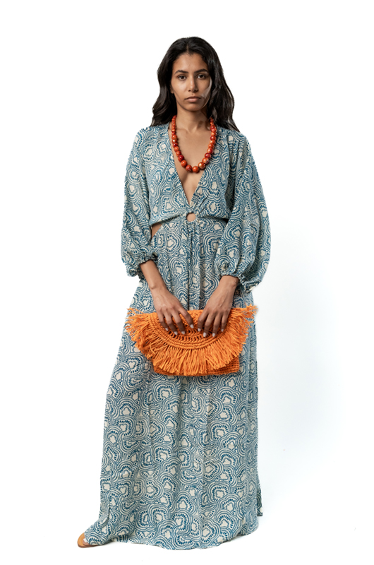 "Model wearing a long blue patterned dress with wide sleeves and a ring detail at the waist, holding an orange fringed clutch and accessorized with a chunky orange necklace, front view."