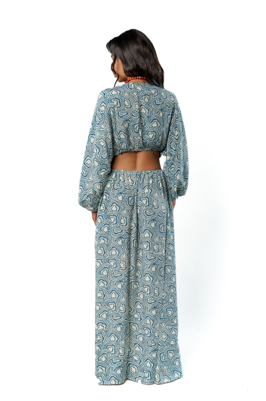 "Back view of a model wearing a long blue patterned dress with wide sleeves, a cutout back design, and an orange necklace, standing with an orange fringed clutch."