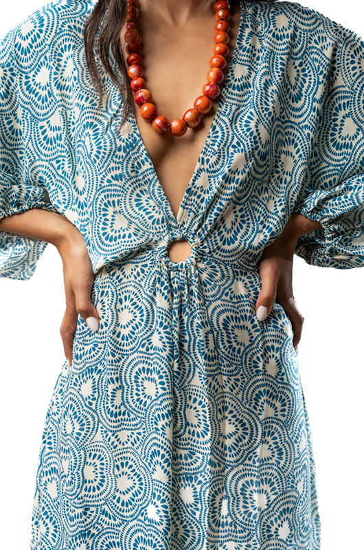 "Close-up of the front waist section of a long blue patterned dress with a ring detail, gathered fabric, and a deep V-neckline, accessorized with a chunky orange necklace."