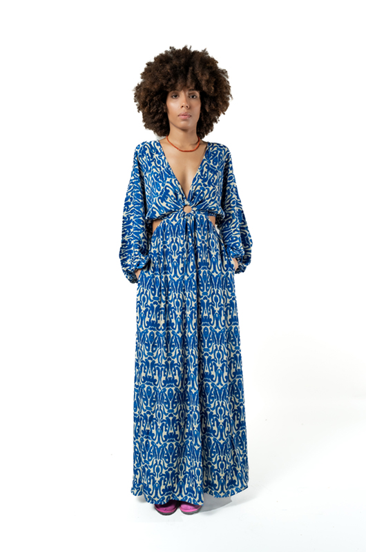 Alt text: Front view of a woman wearing a long-sleeved, deep V-neck blue ikat print maxi dress with a ring detail at the waist and side cutouts.