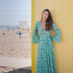 A woman in a green patterned wrap dress with wide sleeves stands barefoot on the beach next to a yellow tiled wall.