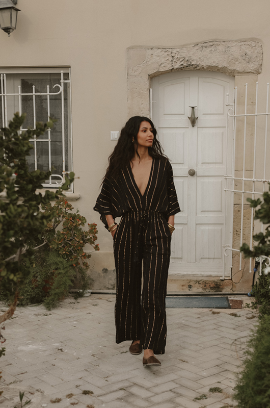 A woman walking outdoors wearing a black wide-leg jumpsuit with gold vertical stripes, a deep V-neckline, and half sleeves, styled casually with flat brown shoes.