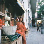 A woman browsing at a market stall while wearing a rust-colored wide-leg jumpsuit with gold vertical stripes, a deep V-neckline, and half sleeves, styled with flat blue shoes.