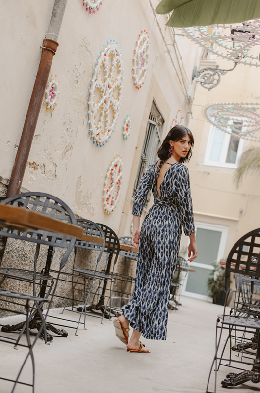 Back view of model walking in a grey curves print jumpsuit with an open back tied at the neck, in a decorated alley.