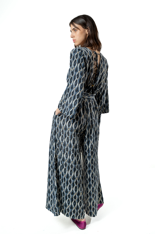 Back three-quarter view of a model wearing a grey curves print jumpsuit with an open back and tied waist, paired with magenta flats.