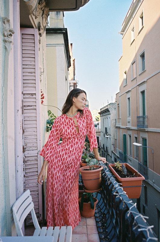 Woman in a pink patterned jumpsuit standing on a balcony with potted plants, looking to the side.