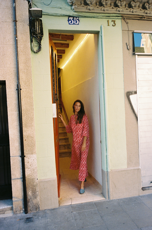 Woman in a pink patterned jumpsuit standing at the entrance of a building with a green facade and wooden stairs inside.