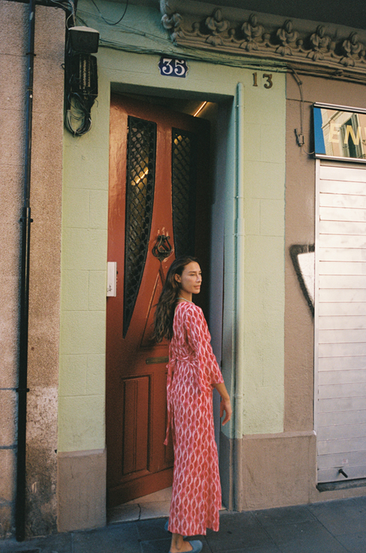 Woman in a pink patterned jumpsuit walking out of a doorway, holding the door open, with the street visible behind her.