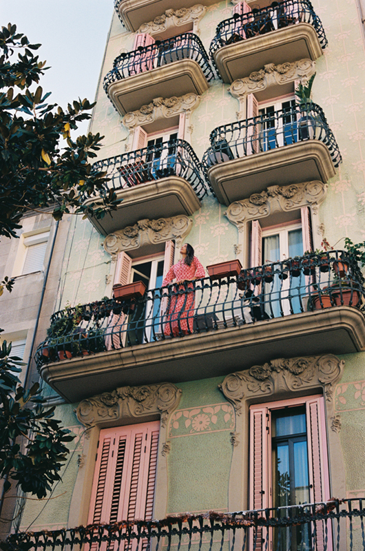 Woman in a pink patterned jumpsuit on the second-floor balcony of an ornate building with green walls and pink shutters.