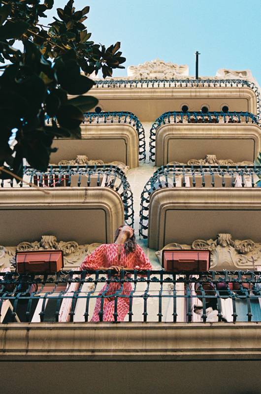 Upward view of a woman in a pink patterned jumpsuit standing on a balcony, looking up at the sky, with iron railings and architectural details in the frame.