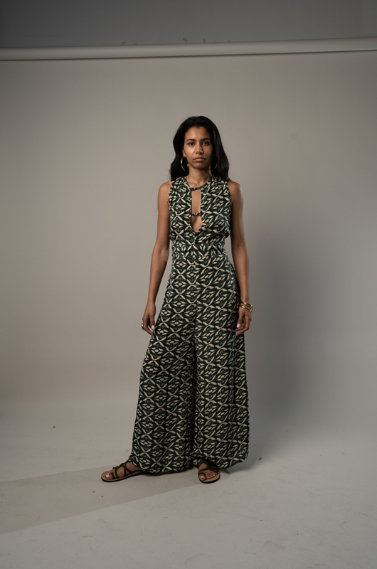 Studio shot of a woman in a green sleeveless printed jumpsuit with a tie-neck design, standing with hands relaxed by her side.