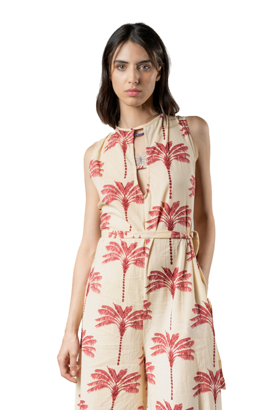 Close-up of a woman in a cream jumpsuit with red palm tree prints, sleeveless with a tie-neck design, looking into the camera.