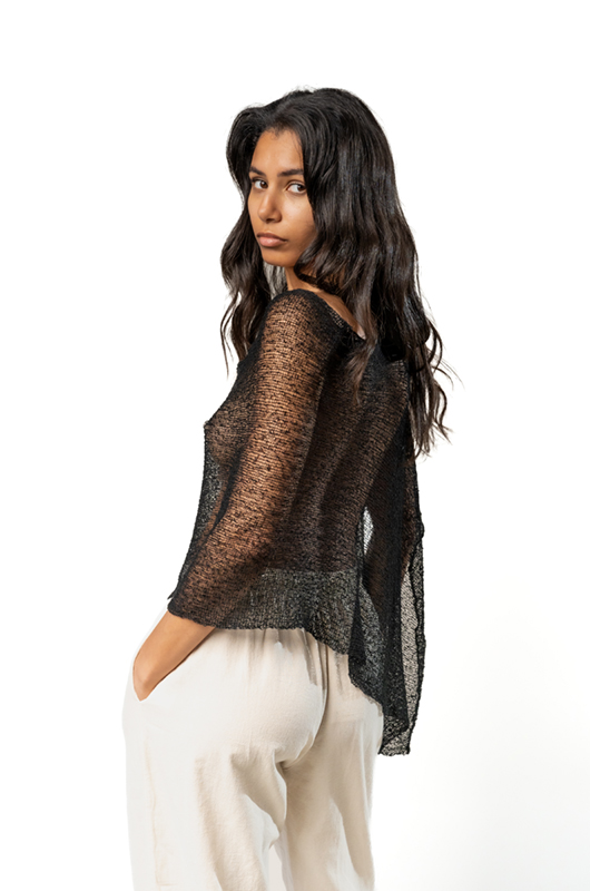 Woman in a sheer black poncho with an asymmetrical hem, paired with cream wide-leg pants, looking over her shoulder.