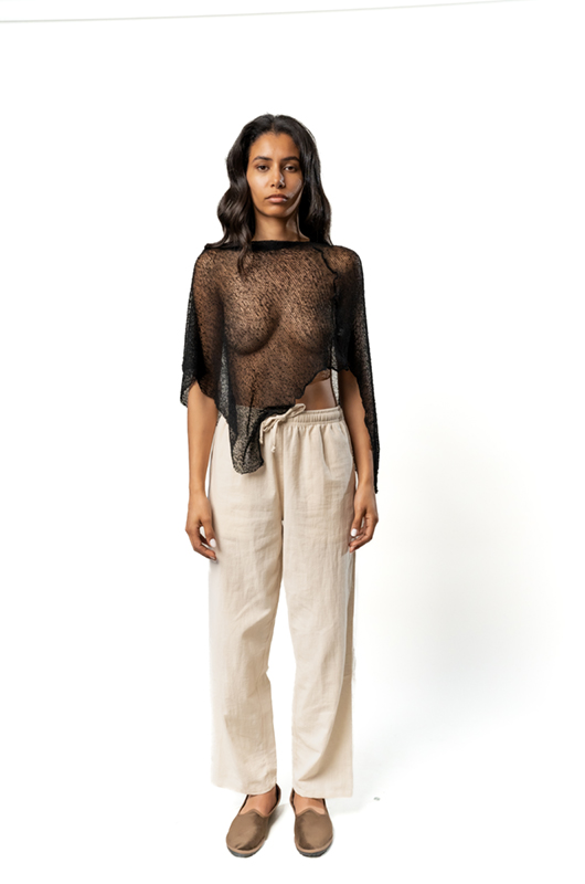 Front view of a sheer black poncho with an asymmetrical hem, worn over cream wide-leg pants.