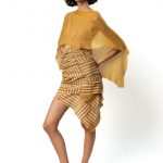 A model is standing sideways wearing a mustard-yellow textured poncho paired with an asymmetrical striped mustard and white skirt, complemented with black sandals decorated with yellow beads.