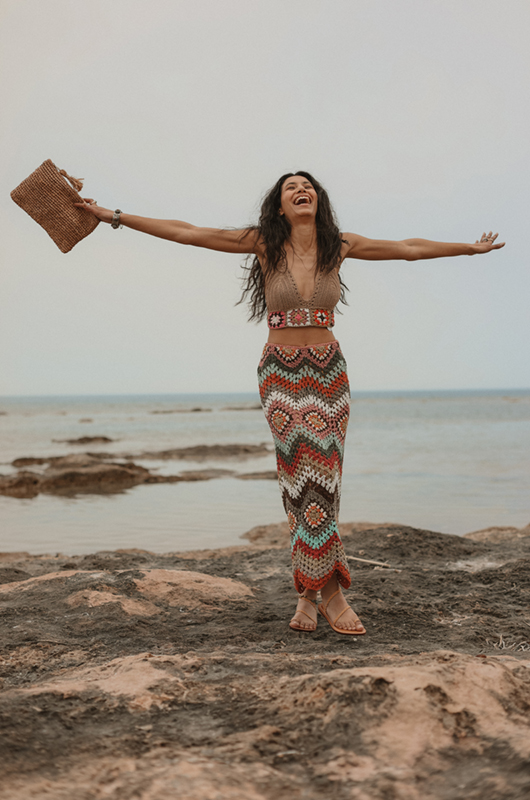 Model wearing a set of multicolor crochet midi skirt in sunset tones and a crochet crop top by rocky shoreline, front view