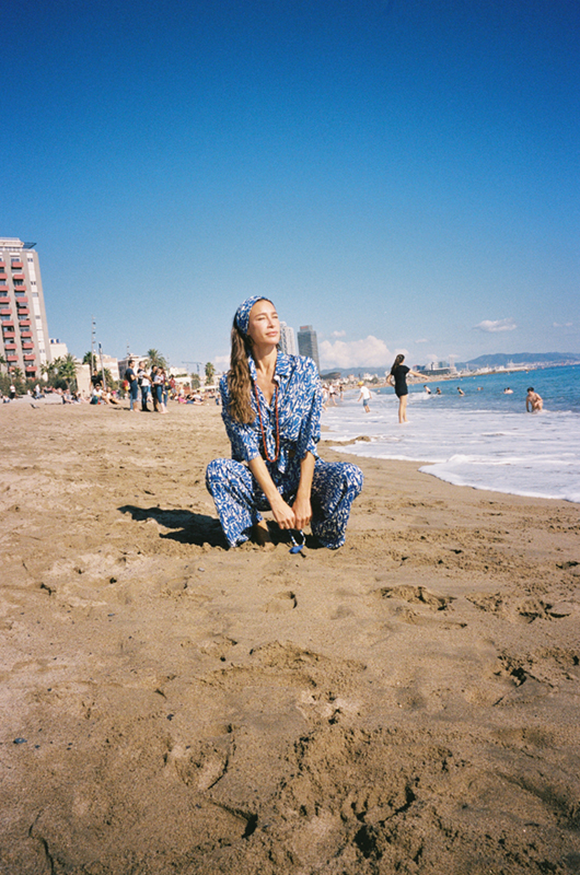 Woman on a beach squatting on the sand, wearing a matching blue and white printed co-ord set with a headband, city skyline in the background.