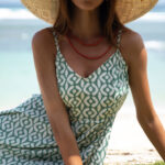 Woman wearing a green and white patterned summer dress with thin straps, accessorized with a straw hat and red necklace, sitting on the grass by the beach with ocean waves in the background