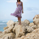 Model standing on seaside rocks wearing a sleeveless V-neck purple and green printed midi dress with a flowy skirt."