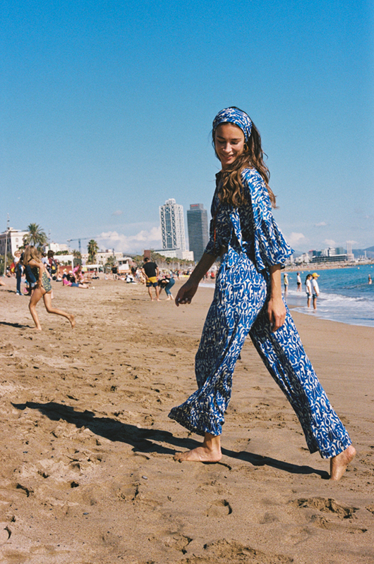 Woman walking barefoot on a sandy beach in a blue and white co-ord set with a headband, smiling as she looks down.