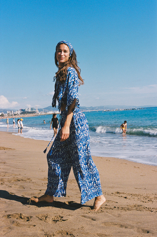 Woman walking along the beach in a blue and white printed co-ord set with a headband, ocean waves and skyline in the background.