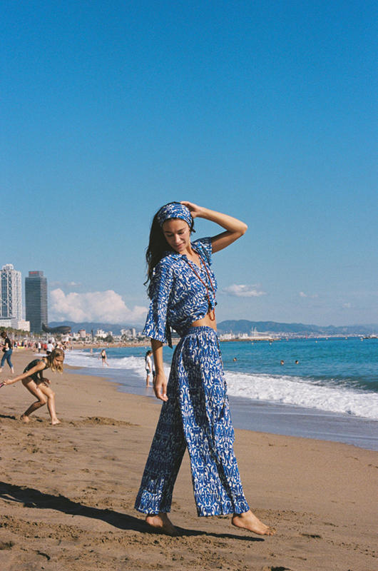 Woman walking along the beach in a blue and white printed co-ord set with a headband, ocean waves and skyline in the background.