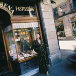 "Street-style photo of a woman leaning outside a bakery window, wearing a dark green and gold printed kimono-style wrap top with a belt and a matching long skirt."