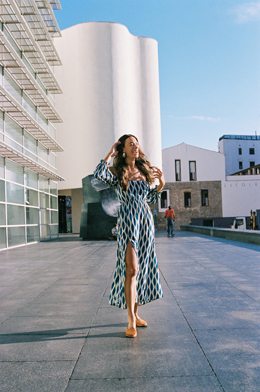 Woman in a long blue off-shoulder geometric print dress walking outdoors in an urban setting