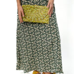 Woman in a black and beige patterned cropped top and matching maxi skirt, holding a small yellow beaded clutch in front, wearing green shoes.