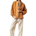 Woman wearing a loose-fit orange patchwork kimono jacket with wide sleeves, ivory trousers, and a chunky black-and-white wooden necklace—posed frontally, holding the jacket closed.