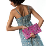Woman in a blue and white patterned strappy dress, holding a bright pink woven clutch behind her back, with short curly hair.