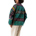 Model facing away wearing a patchwork quilted jacket with geometric and floral patterns in turquoise, purple, and yellow tones, paired with loose beige trousers.